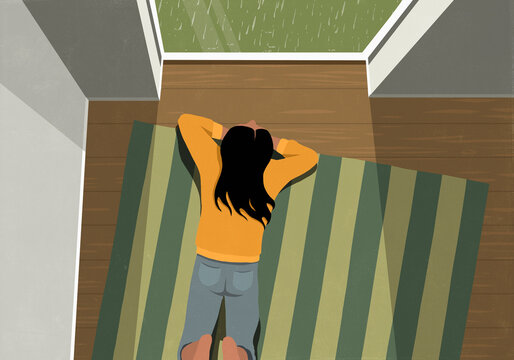View from above of young woman laying on rug and watching rain from window at home