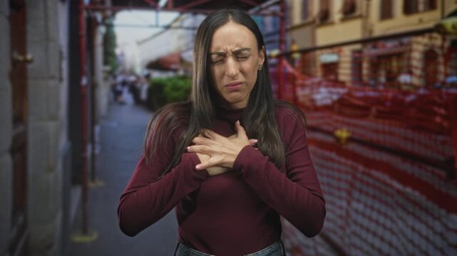 Young hispanic woman clutching chest and grimacing on a city street beside scaffolding and orange construction fence; distress emergency.