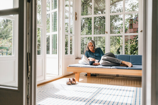 Woman lying on lounge in winter garden reading e-book