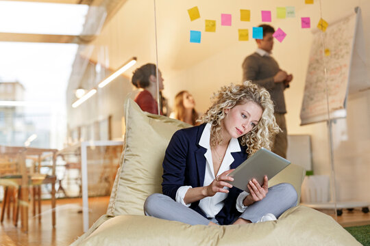 Woman in office using tablet in bean bag with meeting in background