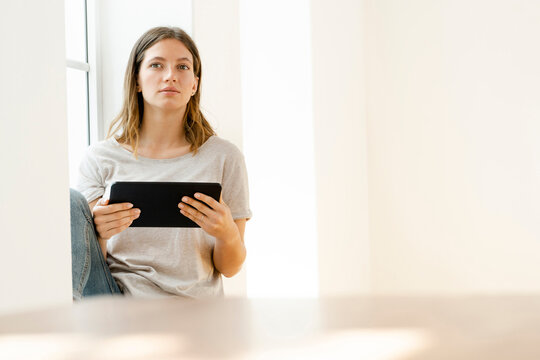 Serious young brunette woman at home sitting on window bench and holding her tablet