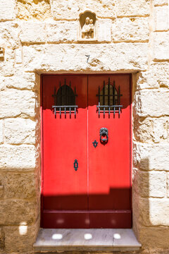Traditional red wooden door with wrought iron details set in a historic limestone facade in the ancient fortified city of Mdina, Malta. Architectural landmark with Mediterranean heritage and timeless 