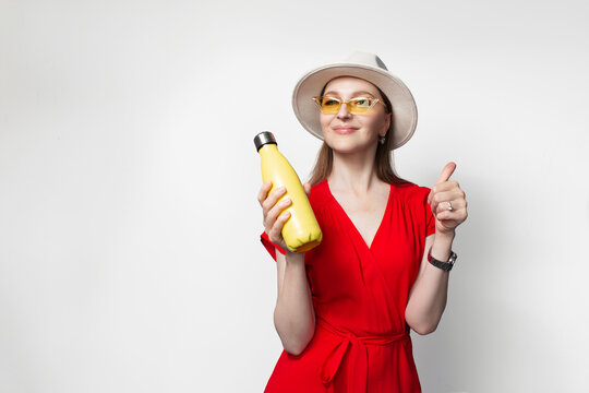 Young girl in red dress wearing glasses and hat, holding thermo bottle in hand, showing thumb up.