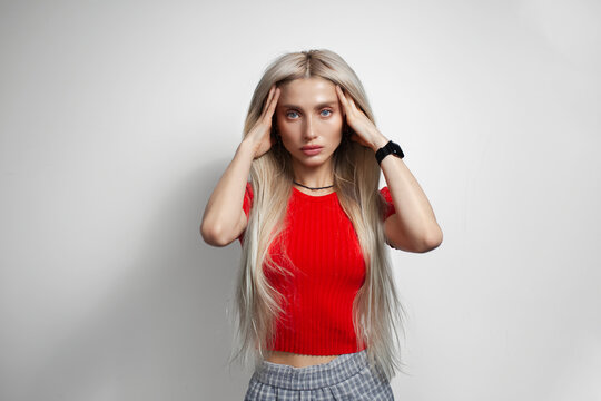 Young pretty blonde girl in red shirt have headache, holding hands on head, on grey background.