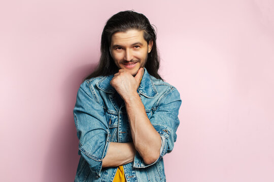 Young happy man with long hair holding hand under chin. Isolated on pastel pink background.