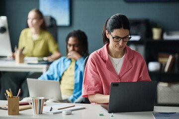 Fototapeta premium Caucasian young adult woman working on laptop in shared office, focusing on task while colleagues using computers in background. Ideal for teamwork, productivity, workplace content