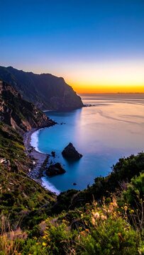 Aerial view of a coastline at dawn. Golden sunlight illuminates a calm sea and beach below rugged cliffs covered in greenery