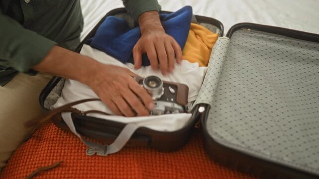 Man packing suitcase with vintage camera and sunglasses, hands securing straps on bed inside building; anticipation travel.