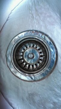 Water Flowing Into Sink Drain. Clear water swirls across a stainless steel basin and drains through a metal strainer.