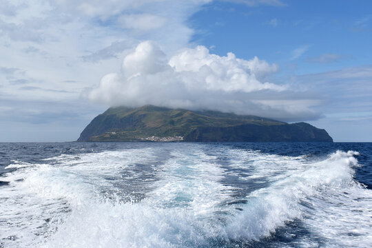 The green island of Corvo partially shrouded in low clouds, with a strong ship's wake churning the ocean surface, Azores