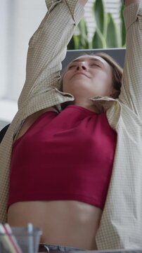 Vertical medium shot of young adult Caucasian woman stretching arms at office desk, then resuming typing on computer. Footage perfect for corporate wellness and software development campaigns