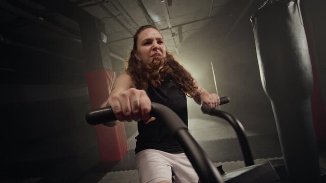 Caucasian young woman training intensely on air resistance exercise bike in dark gym. Suitable for fitness center promotions and motivational athletic marketing campaigns