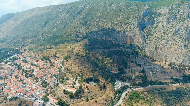 Delphi, Greece. Drone footage of ancient theater and Temple of Apollo ruins on Mount Parnassus slopes, modern town with terracotta rooftops in the valley below, bright summer sun. Drone footage, Poin