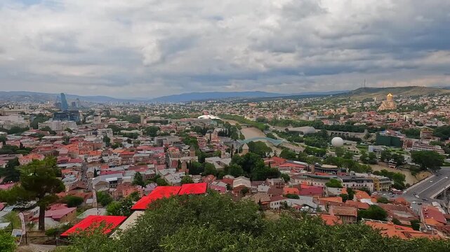 An elevated panorama presents Tbilisi beneath dramatic clouds, with red roofs, the Kura River, Rike Park, historic districts, and cable transport supporting connected tourism and urban development