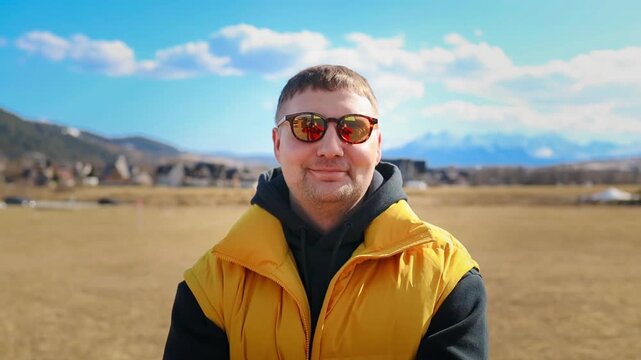 portrait of a happy young caucasian man wearing sunglasses and a bright yellow vest, smiling contentedly while looking away. In the background, a beautiful rural landscape with green hills and village
