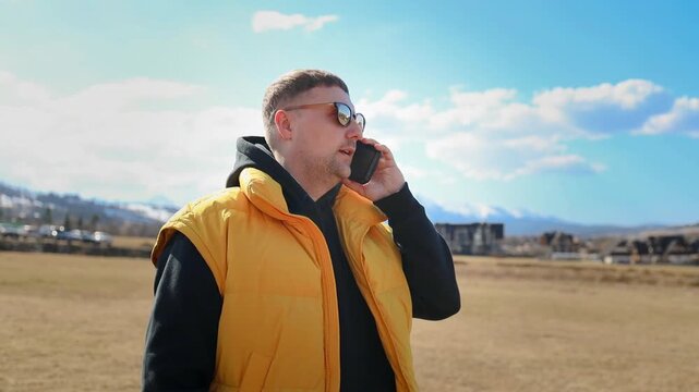 A young caucasian male tourist wearing a yellow vest and sunglasses stands outdoors, talking on his smartphone. He is having a telephone conversation, calling friends, or using roaming services
