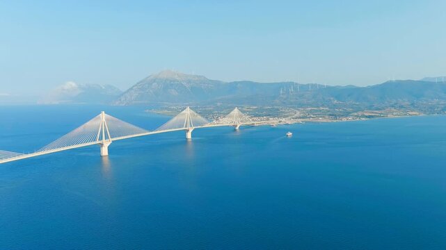 Patras, Greece. The Rio-Antirrio Bridge, a multi-span cable-stayed structure with four white pylons spanning the deep blue Gulf of Corinth, aerial panoramic view under clear summer sky. Drone footage