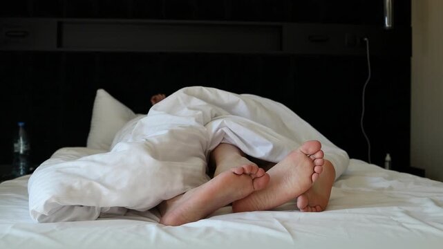 Close up view of a romantic young couple's bare feet crossing and touching together under a soft white blanket or duvet in bed. Captures an intimate, cozy moment of love, connection, tenderness, and