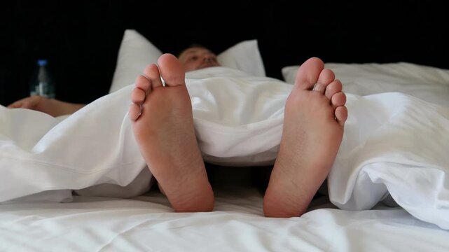 Close up view of bare male feet peaking out from under a soft white blanket or duvet on a bed. In the blurred background, a young man is seen lying down and resting, capturing a comfortable sleep