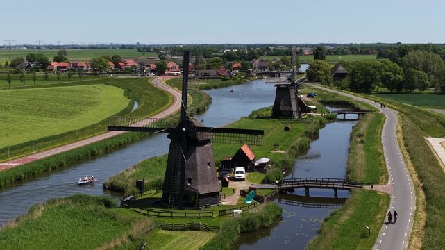 Idyllic aerial perspective of historic windmills lining a picturesque canal in the dutch countryside. With a boat sailing and people walking on a sunny day