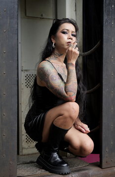 Alternative Asian woman with tattoos crouches in a train doorway while smoking a cigarette