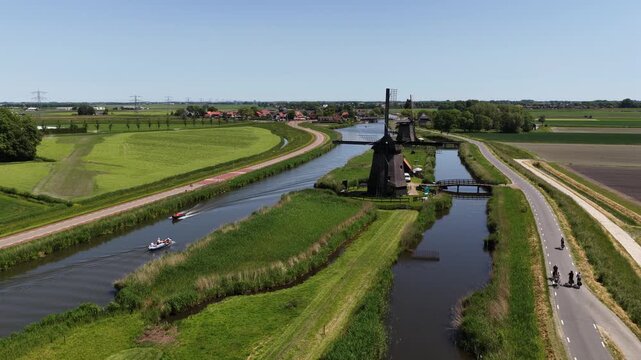 Aerial view of a typical dutch landscape with cyclists riding on a road alongside a canal, a small boat navigating the water, and historic windmills standing among the green polder fields