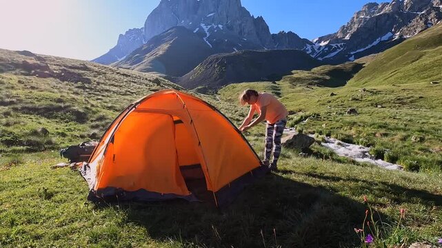 Female camper adjusts a bright orange tent beside an alpine stream, presenting practical shelter setup, self-reliant travel, outdoor accommodation, and expedition readiness in Georgias Caucasus