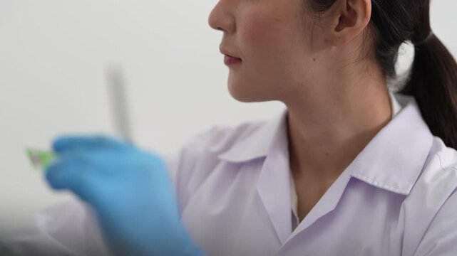 Young Asian woman scientist in white coat and blue gloves lifts a green specimen tube to inspect it in a bright modern laboratory workspace setting