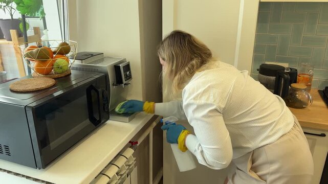 Woman in rubber gloves wiping coffee machine with cloth and holding spray bottle on kitchen
