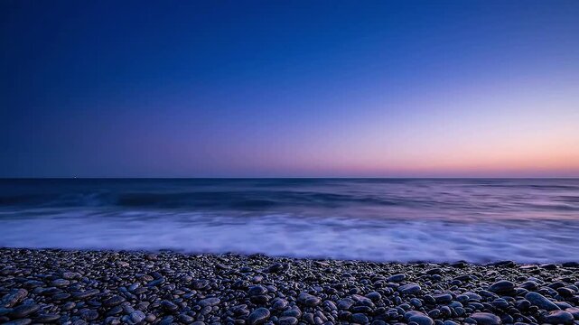 Ocean waves gently wash over a rocky beach at twilight, with a gradient sky transitioning from deep blue to soft orange near the horizon