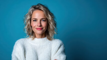 Fototapeta premium A confident woman with short, wavy hair stands against a serene blue backdrop, radiating warmth and positivity in a cozy white sweater, capturing timeless beauty.