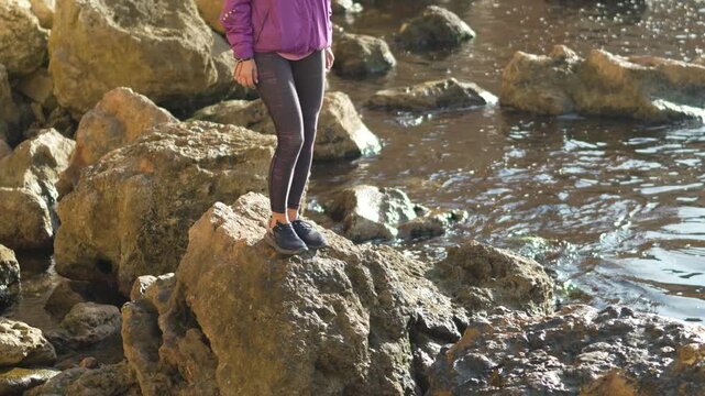 Stepping hiker explores rugged coastal stones by serene sea. Stationary low-angle shot captures precise footwork while transitioning across dry rock formations.