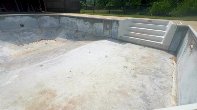 Empty in-ground swimming pool undergoing repair with the vinyl liner removed, exposing the bare concrete steel shell and steps, home renovation and pool maintenance. Panning footage.