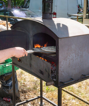 A thin flatbread topped with sliced onion and melted cheese is placed into a portable wood fired metal oven with a long handled peel. Visible flames capture artisan baking, outdoor catering, street fo