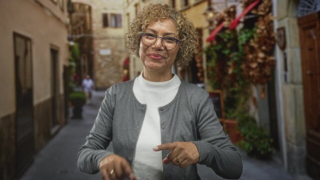 Woman points finger holding keys toward camera on a narrow cobblestone street, smiling and making eye contact; warmth invitation.