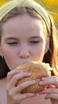 Young girl with a yellow headband eating a fresh hamburger with great appetite in a field. Close up shot showing her enjoying every bite of the fast food meal during an outdoor picnic on a sunny day