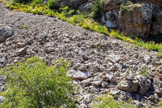 A steep mountain slope covered with loose rocks and scree, bordered by green vegetation and rugged cliffs under bright sunlight.
