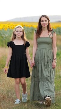 Young woman and her preteen daughter are holding hands and walking towards the camera through a vibrant green field with a beautiful sunflower background during a warm summer day