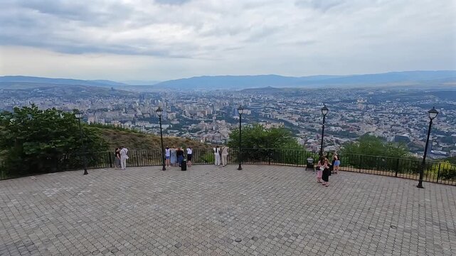 Visitors viewing Tbilisi from elevated overlook above a paved terrace, with the Georgian capital extending toward surrounding hills for tourism promotion