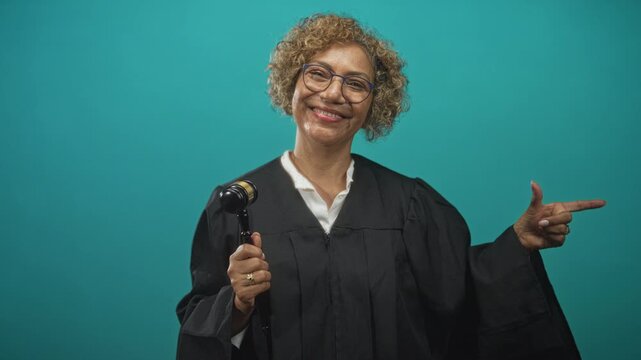 Woman judge in black robe holding gavel and pointing finger to the side in studio setting with teal backdrop, smiling and gesturing; justice confidence.