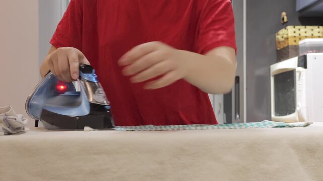 Hands use a steam iron to smooth a checked towel on an ironing board in a home kitchen.