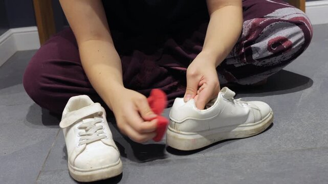 A person wipes the sole edge of a white sneaker with a red sponge on a gray tile floor.