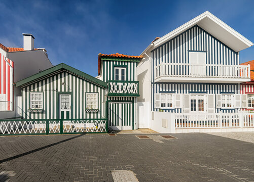 Traditional striped houses with wooden facades, balconies and shutters stand beneath a clear blue sky in Costa Nova, Portugal.