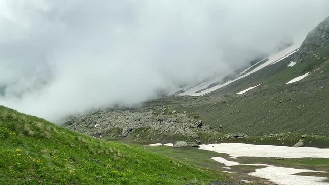 Fog rolling across an alpine slope covers rocky terrain and lingering snow near Chaukhi Pass in Georgia, presenting rapid mountain weather, seasonal route conditions and Caucasus climate geography.