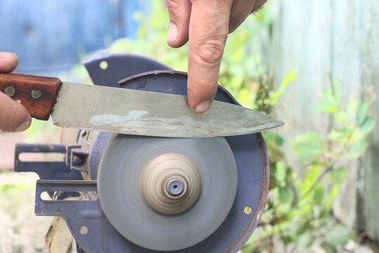 One male worker holds a knife in his hands and sharpens the iron blade on a grinding wheel on an electric sharpening machine on a table