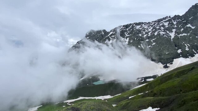 Low clouds sweep across snow-covered Chaukhi slopes above White Abudelauri Lake in Georgia, revealing fragments of turquoise water and creating an alpine weather scene shaped by elevation
