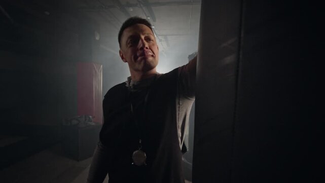 Middle aged Caucasian male boxing coach walking to punching bag, smiling, and practicing punches in dark gym. Perfect for fitness center advertisements and motivational sports promos