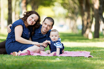 Fototapeta premium Portrait of a smiling family with a baby sitting on a blanket on the green grass in a park on a sunny day.