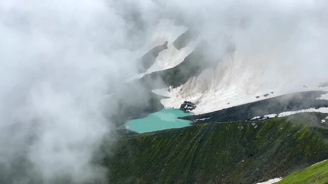 White Abudelauri Lake appears through thick cloud beneath lingering snowfields in Georgia's Caucasus, exposing turquoise meltwater and a severe alpine basin shaped by snowmelt, erosion