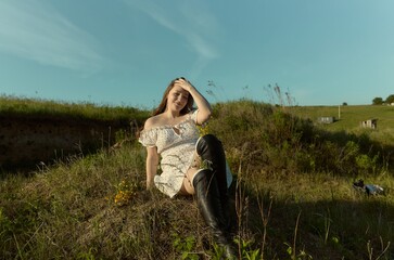 Obraz premium Model sits on a hill covered in grass and flowers while wearing a white dress and black boots during sunny weather
